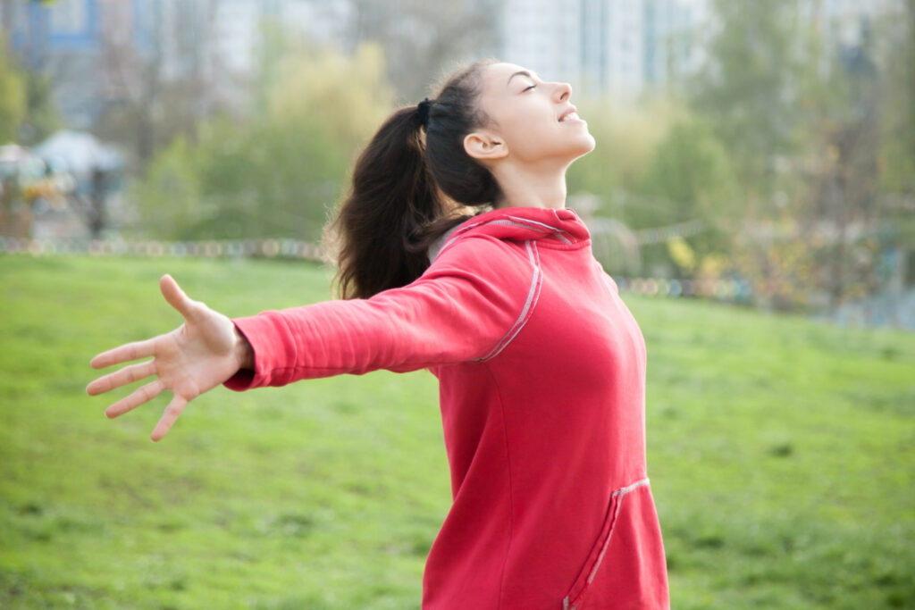 Een vrouw met een rode trui aan, staat op een grasveld. Ze heeft haar armen uitgestrekt. Ze oefent met zang- en ademhalingstechnieken.